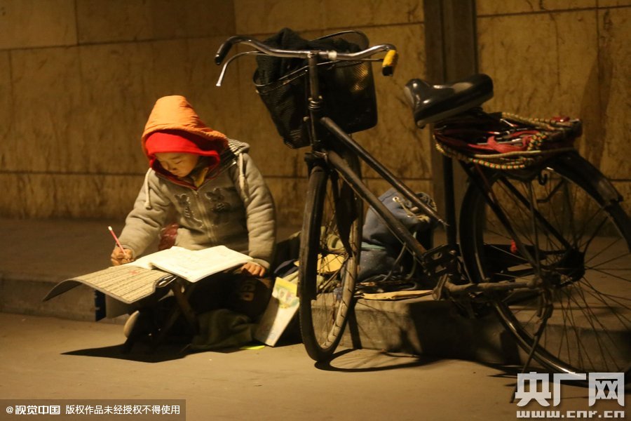 鄭州中原路京廣鐵路大橋下涵洞的馬路牙子邊，一位戴著紅色辣椒帽的小男孩趴在一個(gè)小馬扎上，上面鋪著一張車墊，在微弱的燈光下寫(xiě)作業(yè)